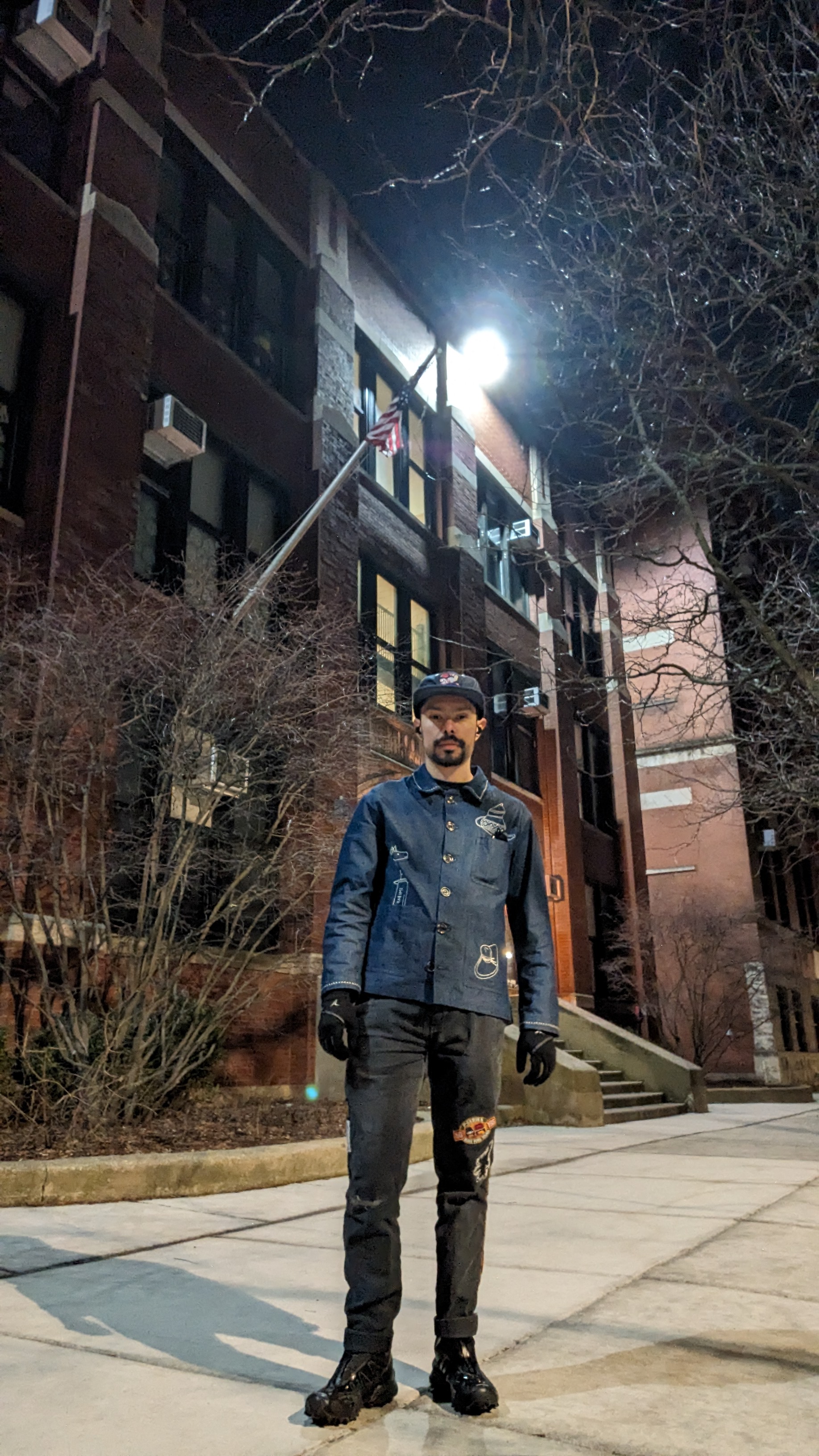 Tyler at night in front of a brick building, wearing a denim jacket and gloves.